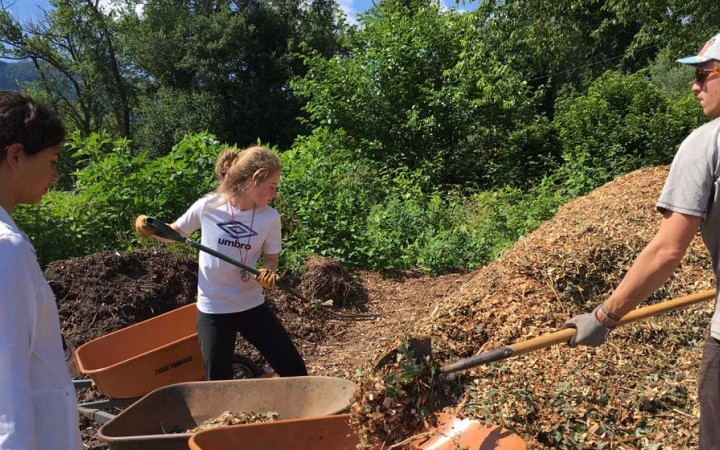 Two people shovel mulch into a wheelbarrow, held by another. 